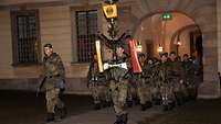 Mehrere Soldaten marschieren in einer Formation durch ein Tor. Sie werden durch Militärmusiker angeführt.