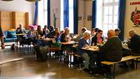 In einem großen Raum sitzen mehrere Menschen an Tischen und essen gemeinsam.