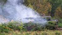 Auf einer Waldlichtung neben Büschen steht ein Panzer im weißen Rauch und schießt.