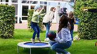 Ein Kind hüpft auf einem Trampolin, eine Frau hockt davor und hält einen Ball. 