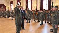 Zwei Soldaten stehen sich gegenüber und grüßen militärisch, ein Zivilist steht daneben.