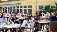 Mehrere Menschen sitzen draußen an Tischen und geniessen ihr Mittagessen.