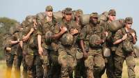 Eine Gruppe von Soldaten marschiert dicht hintereinander mit schwerem Gepäck in sommerlicher Hitze.