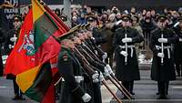 Soldaten in dunklen Militärmänteln mit Truppenfahnen und Nationalflaggen marschieren nebeneinander.