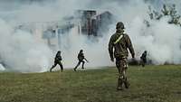 A soldier stands on a green field. In the background, three soldiers run towards a burning building.
