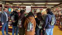 Sieben Personen stehen in einer Bibliothek vor einem Bücherregal und unterhalten sich.