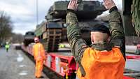 Panzer werden auf einen Zug geladen. Im Vordergrund gibt ein Soldat Handzeichen.