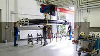 Halle mit Deckenkran, daran hängend ein Torpedo, der von Mitarbeitenden umgelagert wird. 