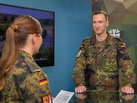 Eine Soldatin interviewt einen Soldaten vor einem Monitor.