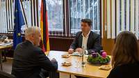 Zwei Männer und eine Frau sitzen sich am Tisch gegenüber.
