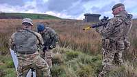 Drei Soldaten stehen auf einer Wiese. Einer davon richtet seine Waffe auf einen mit blauer Mütze.