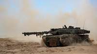 Ein getarnter Kampfpanzer fährt über sandiges Gelände, Staub wirbelt auf.