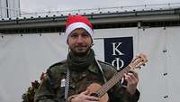 Ein Soldat mit Weihnachtsmütze hält eine kleine Gitarre in der Hand. Im Hintergrund steht ein Gebäude