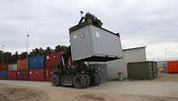 Ein Gabelstapler hebt einen Standardcontainer an, im Hintergrund weitere Container. 