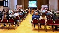 Ein Soldat steht in einem Saal und spricht zu einer Gruppe Menschen in ein Mikrofon.