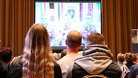 Die Gäste schauen auf eine Leinwand mit einem übertragenen Videobild.
