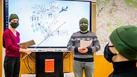 Eine Frau zeigt mit einem Zeigestock auf ein Smartboard auf dem eine Skizze gezeichnet wurde