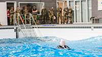 Ein Soldat schwimmt bei Wellengang im Wasser einer Schwimmhalle. Am Beckenrand stehen Soldaten.