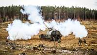 Eine Panzerhaubitze bei der Schussabgabe. Im Hintergrund mit dem nötigem Abstand stehen mehrere Soldaten.