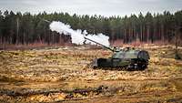 Eine Panzerhaubitze steht auf einem Feld auf dem Truppenübungsplatz. Die Panzerhaubitze gibt gerade einen Schuss ab.
