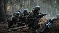 Several soldiers are crouching behind an earthen wall. One of them is holding a weapon and is looking over the wall.