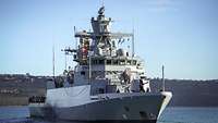 Ein graues Kriegsschiff läuft aus einem Mittelmeerhafen aus.
