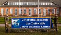 Zwei Soldaten stehen neben einem Schild, welches vor einem Kasernengebäude steht