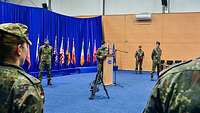Ein Soldat steht an einem Rednerpult. Mehrere Soldatinnen und Soldaten stehen im Vorder und Hintergrund.