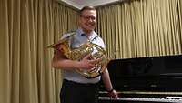 Ein Soldat mit einem Waldhorn in der Hand neben einem Klavier vor einem goldenen Vorhang.