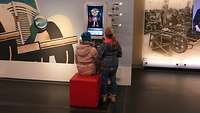 Zwei Kinder stehen an einem Computer und lösen ein Rätsel.