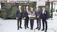 A woman is holding a big key and is standing with four men outside a camouflaged container.