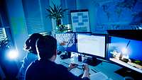 Zwei Personen sitzen in einem abgedunkelten Raum, schauen auf drei Computerbildschirme und analysieren Daten.