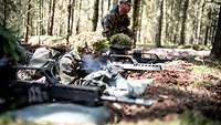 Drei Soldaten liegen in einer Stellung mit dem Gewehr im Anschlag. Im Hintergrund ist der Ausbilder.
