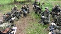 Mehrere Soldaten mit Gewehren und Zetteln in der Hand knien in einem Wald im Kreis.