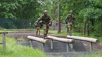 Zwei Soldaten balancieren über die runden Holzbalken, die über einen Graben führen.
