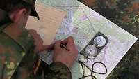 Im Wald hockt ein Soldat am Boden und hält einen Stift, vor sich eine Karte und ein Kompass.