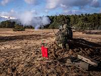Auf einem Feld schießen drei kniende Soldaten mit einer Panzerabwehrwaffe. Rauch entsteht.