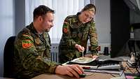Ein Soldat sitzt an einem Schreibtisch, daneben steht eine Soldatin. Sie weist auf Papier vor ihm.
