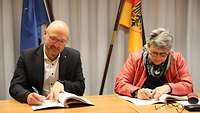 A man and a woman are sitting at a table and are writing on paper. Behind them are two flags.