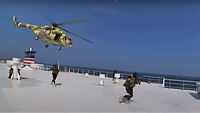 Bewaffnete auf dem Oberdeck eines Schiffs, darüber ein Hubschrauber vor blauem Himmel.