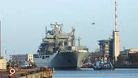 A small blue-and-white boat pulling a large gray ship on the water.