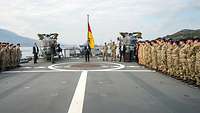 Auf dem Flugdeck eines Kriegsschiffs steht ein Mann im blauen Jackett zwischen zwei Reihen angetretener Soldaten.