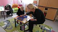 Eine Kinderbetreuerin sitzt mit einem Kind an einem Tisch und spielt. 