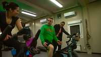 Zwei Soldatinnen und ein Soldat in Sportkleidung sitzen nebeneinander auf Trainingsrädern