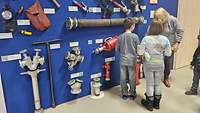 Zwei Kinder stehen vor einer Wand mit Feuerwehrschläuchen.