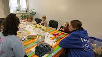 Zwei Kinderbetreuerinnen sitzen mit Kindern an einem Tisch und basteln Faschingsmasken.
