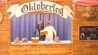 Zwei Angehörige stehen mit Kunstbierkrügen in einem nachgebauten Oktoberfestausschank.