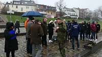Eine Gruppe von Personen steht, teils mit Regenschirm, auf einem Platz.