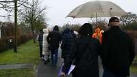 Eine Gruppe Personen geht einen Weg entlang, dabei trägt eine Frau einen Regenschirm.