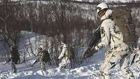 In einem bergigen Gebiet voller Schnee und Sträucher gehen bewaffnete Soldaten einen Hang hinunter